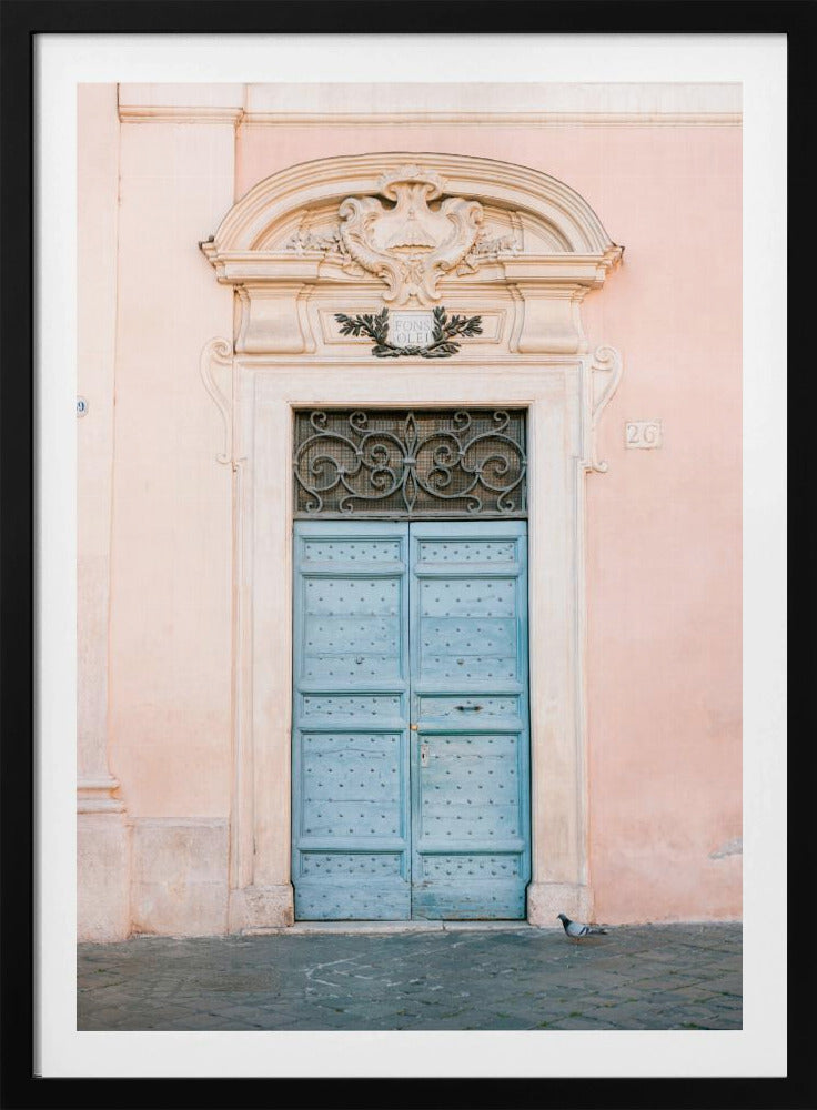 A light blue, studded double door set into a pale pink wall. The doorway is framed in white stone with an ornate pediment and a decorative wrought-iron transom window above the door. A single pigeon stands on the cobblestone pavement in front. Print