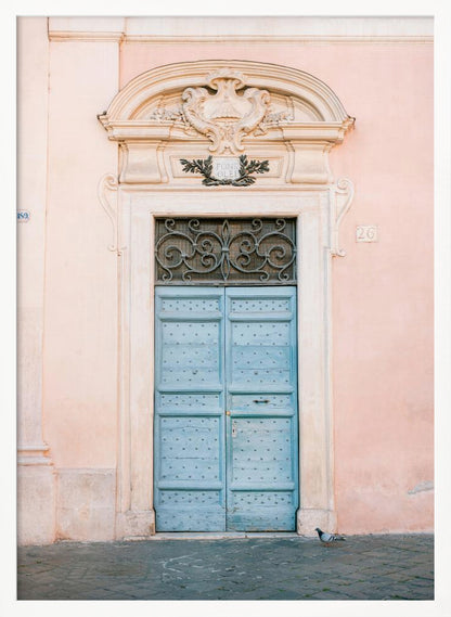 A light blue, studded double door set into a pale pink wall. The doorway is framed in white stone with an ornate pediment and a decorative wrought-iron transom window above the door. A single pigeon stands on the cobblestone pavement in front. Print