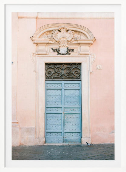 A light blue, studded double door set into a pale pink wall. The doorway is framed in white stone with an ornate pediment and a decorative wrought-iron transom window above the door. A single pigeon stands on the cobblestone pavement in front. Print