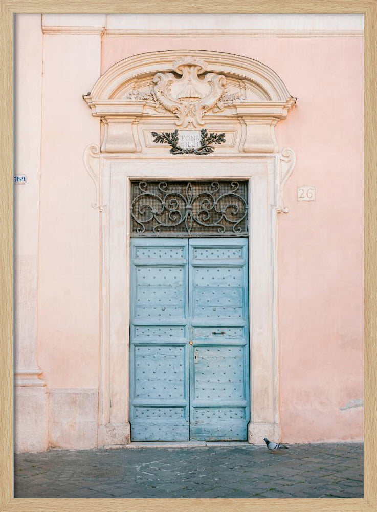 A light blue, studded double door set into a pale pink wall. The doorway is framed in white stone with an ornate pediment and a decorative wrought-iron transom window above the door. A single pigeon stands on the cobblestone pavement in front. Print