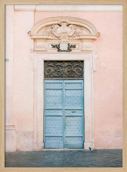 A light blue, studded double door set into a pale pink wall. The doorway is framed in white stone with an ornate pediment and a decorative wrought-iron transom window above the door. A single pigeon stands on the cobblestone pavement in front. Print