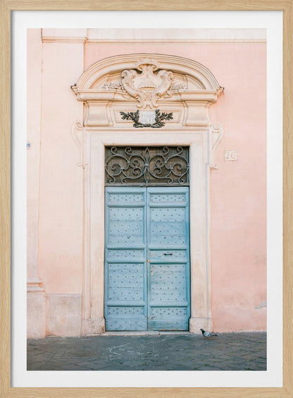 A light blue, studded double door set into a pale pink wall. The doorway is framed in white stone with an ornate pediment and a decorative wrought-iron transom window above the door. A single pigeon stands on the cobblestone pavement in front. Print