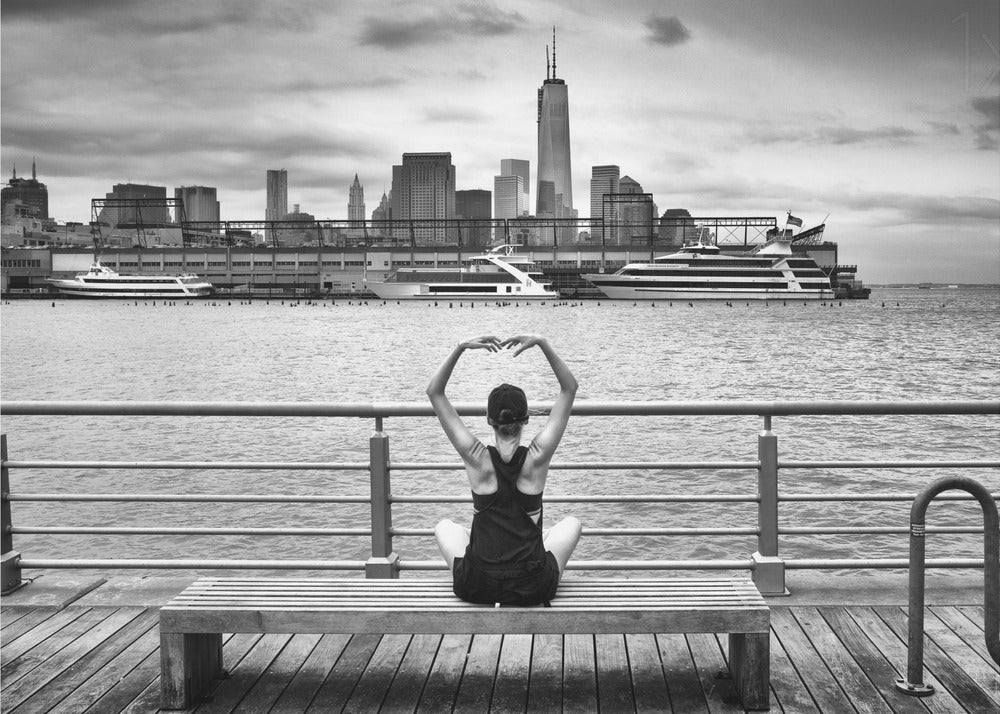 A framed black and white photograph showing the back of a woman sitting on a wooden bench on a pier. She is in a yoga pose with her arms raised, looking out at the Lower Manhattan skyline, including the Freedom Tower, across the water. Decor