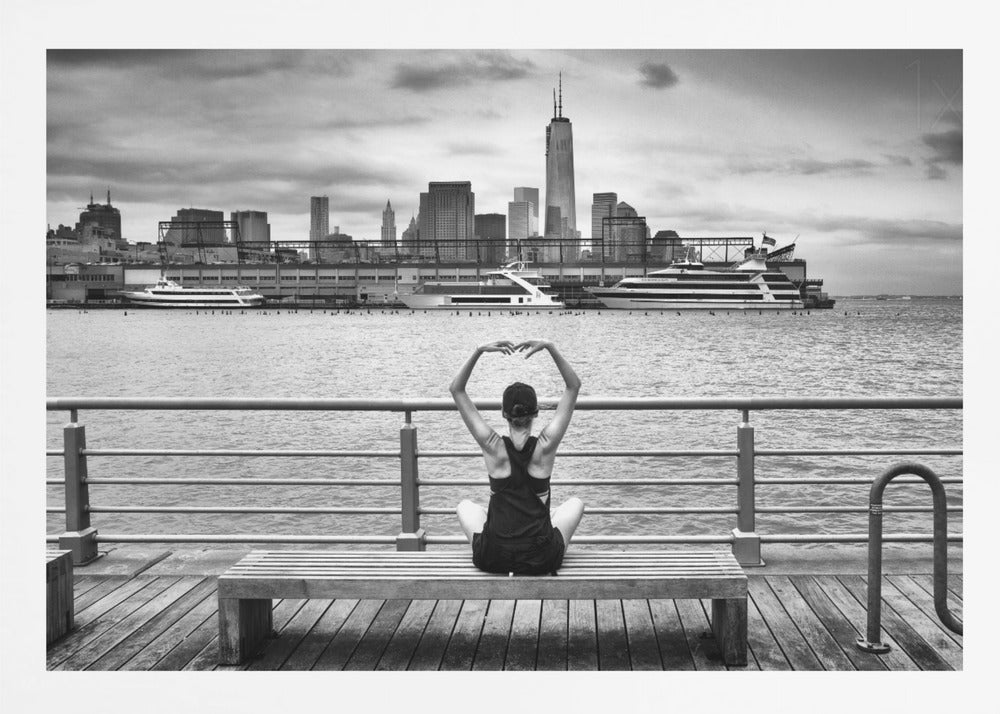 A framed black and white photograph showing the back of a woman sitting on a wooden bench on a pier. She is in a yoga pose with her arms raised, looking out at the Lower Manhattan skyline, including the Freedom Tower, across the water. Decor