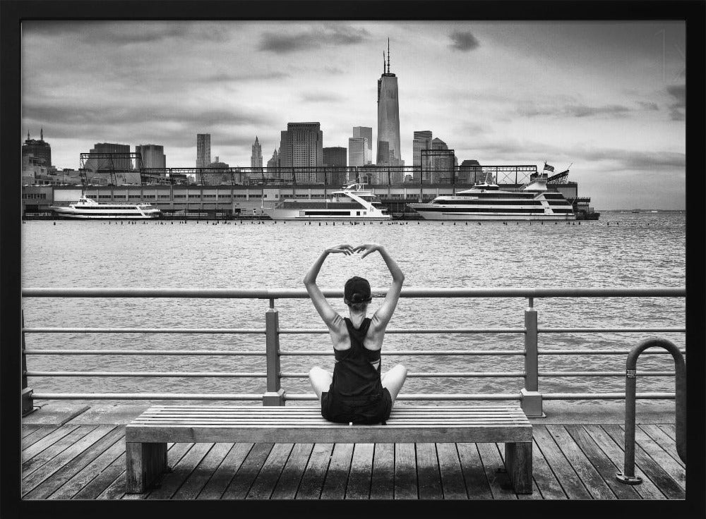 A framed black and white photograph showing the back of a woman sitting on a wooden bench on a pier. She is in a yoga pose with her arms raised, looking out at the Lower Manhattan skyline, including the Freedom Tower, across the water. Decor
