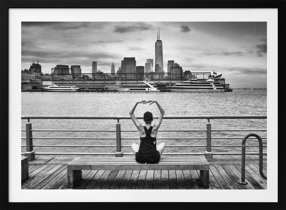 A framed black and white photograph showing the back of a woman sitting on a wooden bench on a pier. She is in a yoga pose with her arms raised, looking out at the Lower Manhattan skyline, including the Freedom Tower, across the water. Decor