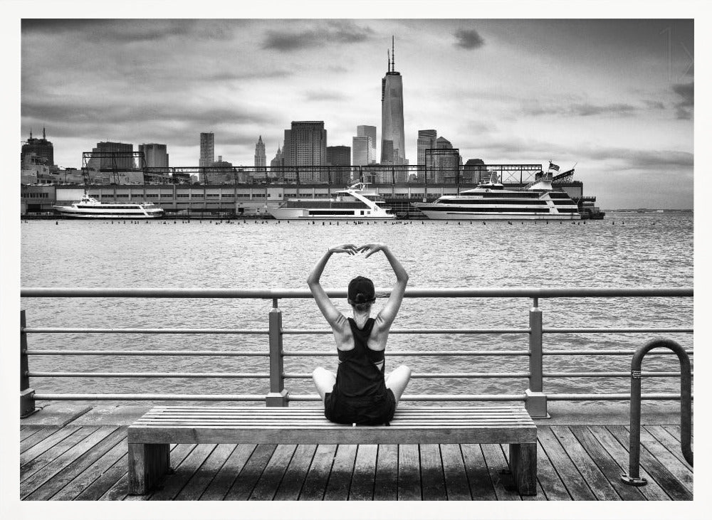 A framed black and white photograph showing the back of a woman sitting on a wooden bench on a pier. She is in a yoga pose with her arms raised, looking out at the Lower Manhattan skyline, including the Freedom Tower, across the water. Decor