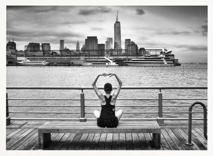 A framed black and white photograph showing the back of a woman sitting on a wooden bench on a pier. She is in a yoga pose with her arms raised, looking out at the Lower Manhattan skyline, including the Freedom Tower, across the water. Decor