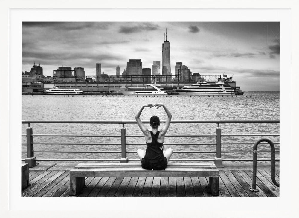 A framed black and white photograph showing the back of a woman sitting on a wooden bench on a pier. She is in a yoga pose with her arms raised, looking out at the Lower Manhattan skyline, including the Freedom Tower, across the water. Decor