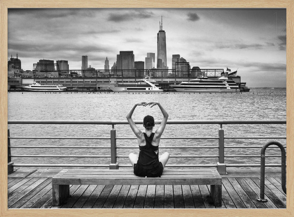 A framed black and white photograph showing the back of a woman sitting on a wooden bench on a pier. She is in a yoga pose with her arms raised, looking out at the Lower Manhattan skyline, including the Freedom Tower, across the water. Decor