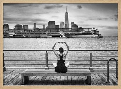 A framed black and white photograph showing the back of a woman sitting on a wooden bench on a pier. She is in a yoga pose with her arms raised, looking out at the Lower Manhattan skyline, including the Freedom Tower, across the water. Decor