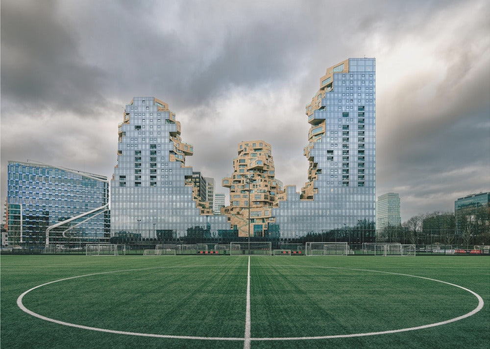 A wide-angle view from the center of a lush green soccer field looking towards a striking modern architectural complex with uniquely shaped glass and tan buildings under a dramatic, cloudy sky. Decor