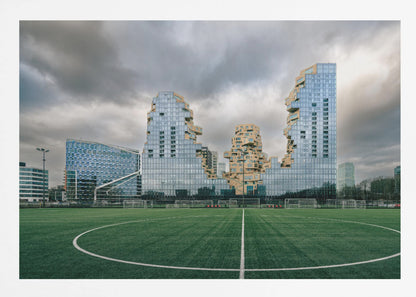 A wide-angle view from the center of a lush green soccer field looking towards a striking modern architectural complex with uniquely shaped glass and tan buildings under a dramatic, cloudy sky. Decor