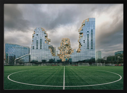 A wide-angle view from the center of a lush green soccer field looking towards a striking modern architectural complex with uniquely shaped glass and tan buildings under a dramatic, cloudy sky. Decor