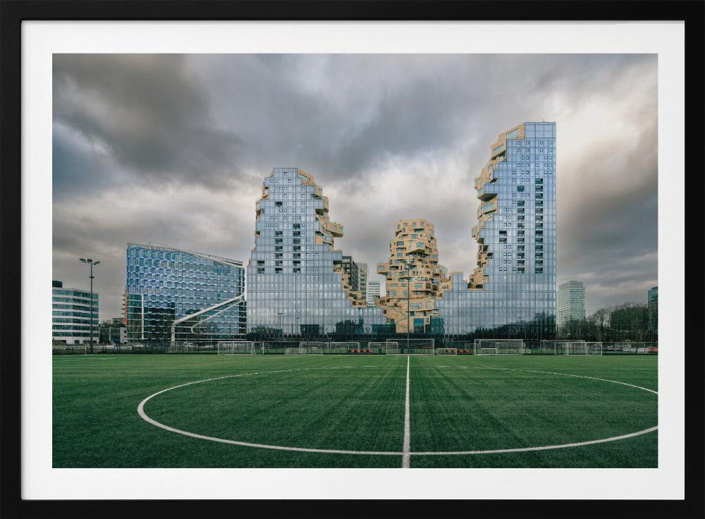 A wide-angle view from the center of a lush green soccer field looking towards a striking modern architectural complex with uniquely shaped glass and tan buildings under a dramatic, cloudy sky. Decor