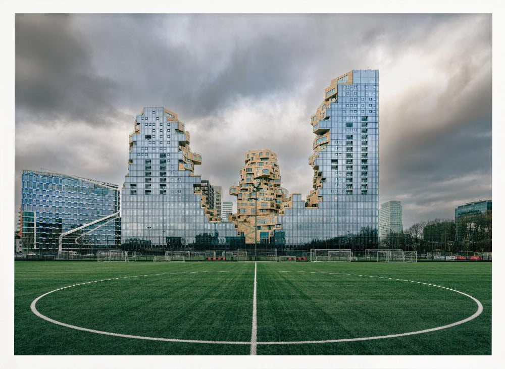 A wide-angle view from the center of a lush green soccer field looking towards a striking modern architectural complex with uniquely shaped glass and tan buildings under a dramatic, cloudy sky. Decor