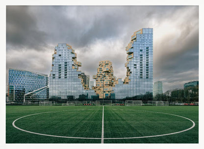 A wide-angle view from the center of a lush green soccer field looking towards a striking modern architectural complex with uniquely shaped glass and tan buildings under a dramatic, cloudy sky. Decor