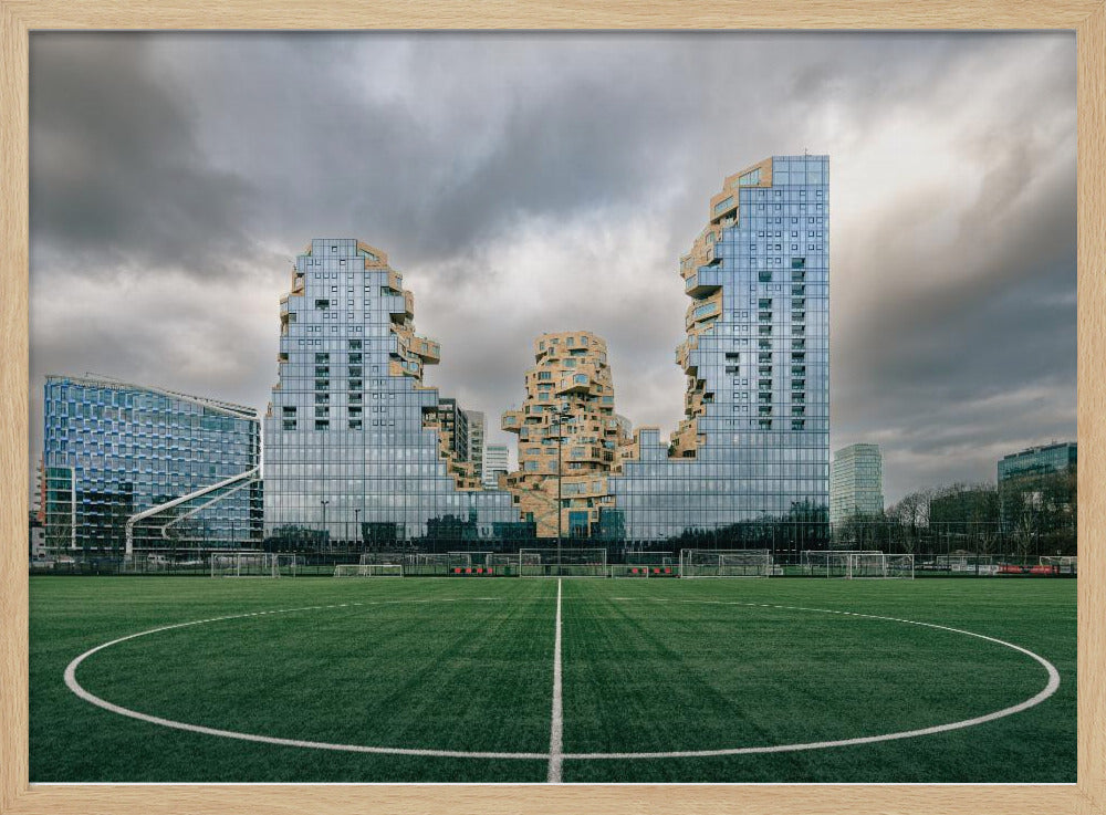 A wide-angle view from the center of a lush green soccer field looking towards a striking modern architectural complex with uniquely shaped glass and tan buildings under a dramatic, cloudy sky. Decor