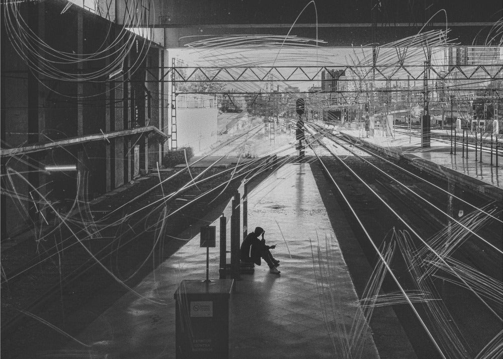 A framed, high-contrast black and white photograph of a person sitting alone on a deserted train platform, with tracks stretching into the distance. The entire scene is overlaid with frenetic white scribbles, creating an abstract and moody effect. Artwork