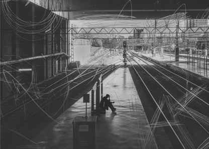A framed, high-contrast black and white photograph of a person sitting alone on a deserted train platform, with tracks stretching into the distance. The entire scene is overlaid with frenetic white scribbles, creating an abstract and moody effect. Artwork