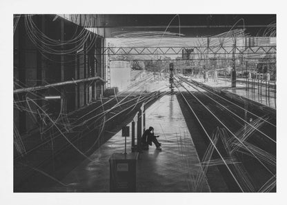 A framed, high-contrast black and white photograph of a person sitting alone on a deserted train platform, with tracks stretching into the distance. The entire scene is overlaid with frenetic white scribbles, creating an abstract and moody effect. Artwork
