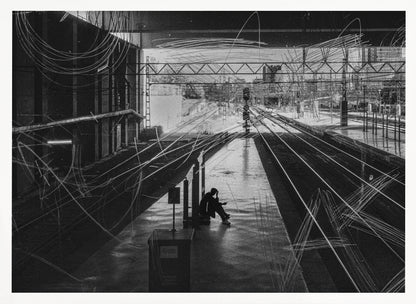 A framed, high-contrast black and white photograph of a person sitting alone on a deserted train platform, with tracks stretching into the distance. The entire scene is overlaid with frenetic white scribbles, creating an abstract and moody effect. Artwork