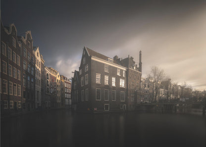 A moody, atmospheric photograph of historic canal houses in Amsterdam, viewed from the water. The scene is dark, with a dramatic, cloudy sky and a soft glow on the horizon, while some windows in the dark brick buildings are lit from within. The image is bordered by a silver frame. Wall Art