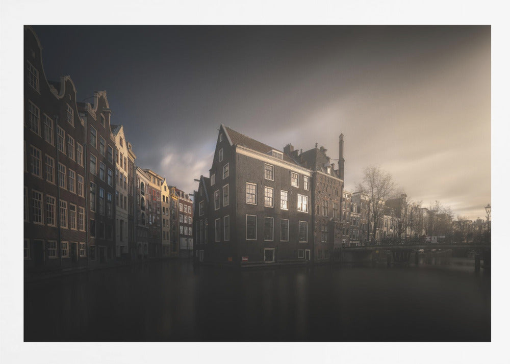 A moody, atmospheric photograph of historic canal houses in Amsterdam, viewed from the water. The scene is dark, with a dramatic, cloudy sky and a soft glow on the horizon, while some windows in the dark brick buildings are lit from within. The image is bordered by a silver frame. Wall Art