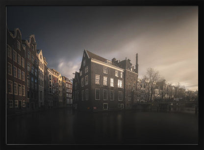A moody, atmospheric photograph of historic canal houses in Amsterdam, viewed from the water. The scene is dark, with a dramatic, cloudy sky and a soft glow on the horizon, while some windows in the dark brick buildings are lit from within. The image is bordered by a silver frame. Wall Art