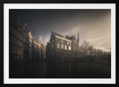 A moody, atmospheric photograph of historic canal houses in Amsterdam, viewed from the water. The scene is dark, with a dramatic, cloudy sky and a soft glow on the horizon, while some windows in the dark brick buildings are lit from within. The image is bordered by a silver frame. Wall Art