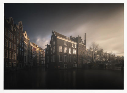 A moody, atmospheric photograph of historic canal houses in Amsterdam, viewed from the water. The scene is dark, with a dramatic, cloudy sky and a soft glow on the horizon, while some windows in the dark brick buildings are lit from within. The image is bordered by a silver frame. Wall Art