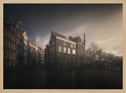A moody, atmospheric photograph of historic canal houses in Amsterdam, viewed from the water. The scene is dark, with a dramatic, cloudy sky and a soft glow on the horizon, while some windows in the dark brick buildings are lit from within. The image is bordered by a silver frame. Wall Art