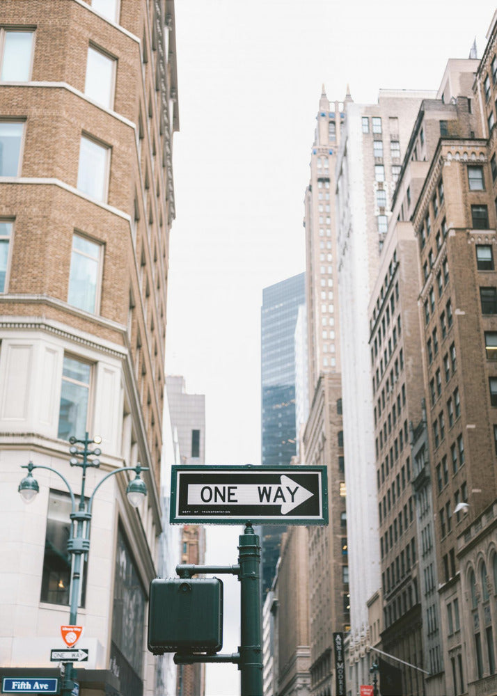 A framed photograph of a New York City street, with a prominent 'ONE WAY' sign in the foreground pointing right. The view is from a low angle, looking up between tall brick and stone buildings towards a bright, overcast sky. Wall Art