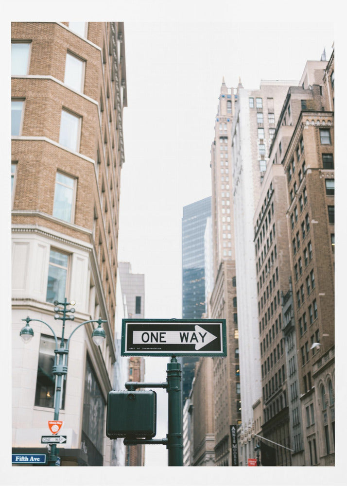 A framed photograph of a New York City street, with a prominent 'ONE WAY' sign in the foreground pointing right. The view is from a low angle, looking up between tall brick and stone buildings towards a bright, overcast sky. Wall Art