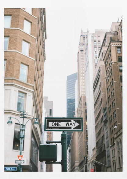 A framed photograph of a New York City street, with a prominent 'ONE WAY' sign in the foreground pointing right. The view is from a low angle, looking up between tall brick and stone buildings towards a bright, overcast sky. Wall Art