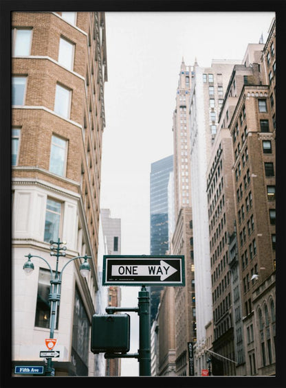 A framed photograph of a New York City street, with a prominent 'ONE WAY' sign in the foreground pointing right. The view is from a low angle, looking up between tall brick and stone buildings towards a bright, overcast sky. Wall Art