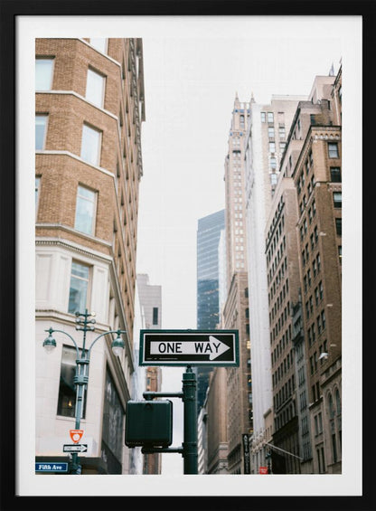A framed photograph of a New York City street, with a prominent 'ONE WAY' sign in the foreground pointing right. The view is from a low angle, looking up between tall brick and stone buildings towards a bright, overcast sky. Wall Art