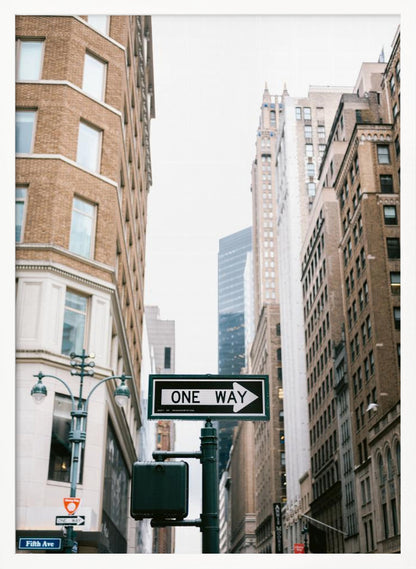 A framed photograph of a New York City street, with a prominent 'ONE WAY' sign in the foreground pointing right. The view is from a low angle, looking up between tall brick and stone buildings towards a bright, overcast sky. Wall Art