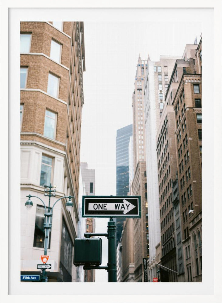 A framed photograph of a New York City street, with a prominent 'ONE WAY' sign in the foreground pointing right. The view is from a low angle, looking up between tall brick and stone buildings towards a bright, overcast sky. Wall Art