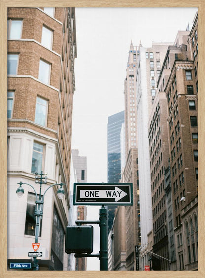 A framed photograph of a New York City street, with a prominent 'ONE WAY' sign in the foreground pointing right. The view is from a low angle, looking up between tall brick and stone buildings towards a bright, overcast sky. Wall Art