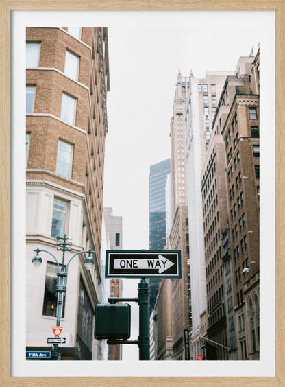 A framed photograph of a New York City street, with a prominent 'ONE WAY' sign in the foreground pointing right. The view is from a low angle, looking up between tall brick and stone buildings towards a bright, overcast sky. Wall Art