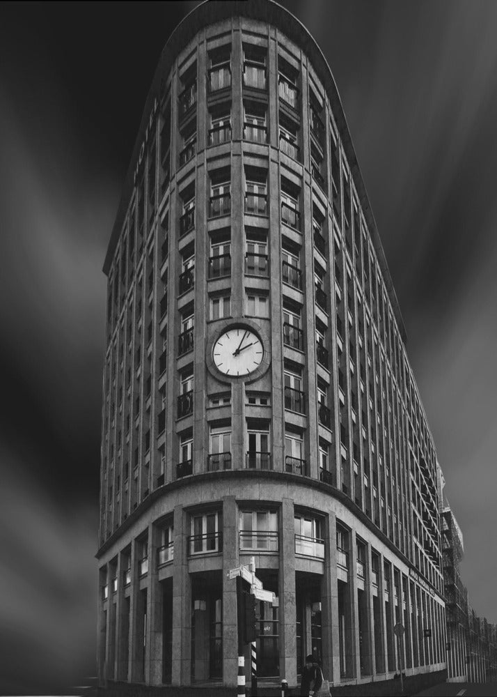 A dramatic low-angle, black and white photograph of a large, curved corner building with a prominent clock on its facade. The sky is dark with a motion blur effect, making the building appear grand and imposing. Artwork