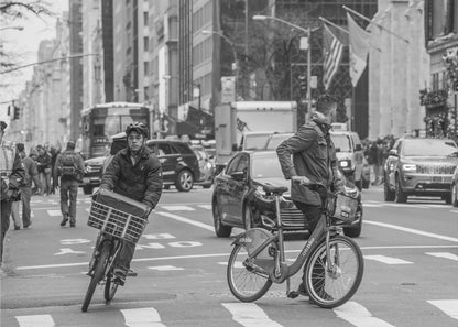 A black and white street photograph of a busy city intersection, framed in silver. In the foreground, two men are crossing with their bicycles amidst traffic, with one cyclist looking directly at the camera. Tall buildings and other pedestrians line the background, capturing a moment of everyday city life. Artwork