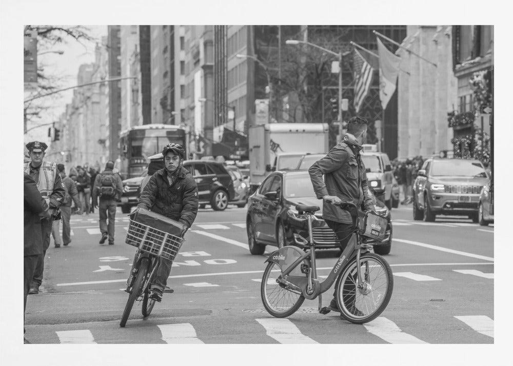 A black and white street photograph of a busy city intersection, framed in silver. In the foreground, two men are crossing with their bicycles amidst traffic, with one cyclist looking directly at the camera. Tall buildings and other pedestrians line the background, capturing a moment of everyday city life. Artwork