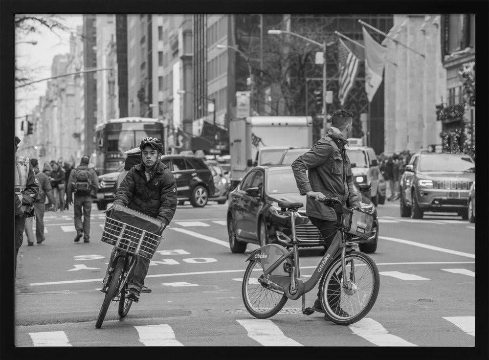 A black and white street photograph of a busy city intersection, framed in silver. In the foreground, two men are crossing with their bicycles amidst traffic, with one cyclist looking directly at the camera. Tall buildings and other pedestrians line the background, capturing a moment of everyday city life. Artwork