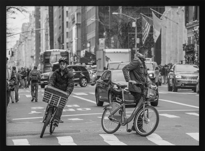 A black and white street photograph of a busy city intersection, framed in silver. In the foreground, two men are crossing with their bicycles amidst traffic, with one cyclist looking directly at the camera. Tall buildings and other pedestrians line the background, capturing a moment of everyday city life. Artwork