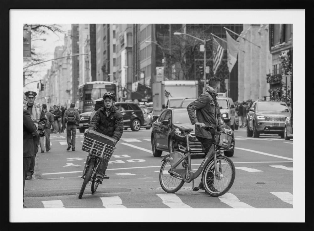 A black and white street photograph of a busy city intersection, framed in silver. In the foreground, two men are crossing with their bicycles amidst traffic, with one cyclist looking directly at the camera. Tall buildings and other pedestrians line the background, capturing a moment of everyday city life. Artwork