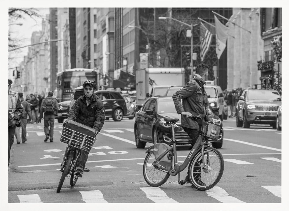 A black and white street photograph of a busy city intersection, framed in silver. In the foreground, two men are crossing with their bicycles amidst traffic, with one cyclist looking directly at the camera. Tall buildings and other pedestrians line the background, capturing a moment of everyday city life. Artwork