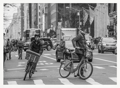 A black and white street photograph of a busy city intersection, framed in silver. In the foreground, two men are crossing with their bicycles amidst traffic, with one cyclist looking directly at the camera. Tall buildings and other pedestrians line the background, capturing a moment of everyday city life. Artwork