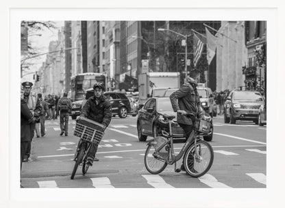 A black and white street photograph of a busy city intersection, framed in silver. In the foreground, two men are crossing with their bicycles amidst traffic, with one cyclist looking directly at the camera. Tall buildings and other pedestrians line the background, capturing a moment of everyday city life. Artwork